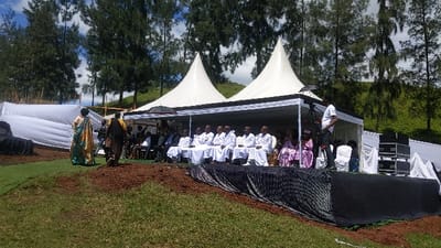 The image shows a scene in Rukoko, Democratic Republic of Congo (latitude: -1.624, longitude: 29.258). There are two white tents with black accents set up on a grassy slope. Under the tents, a group of men in white robes are seated in chairs, facing the camera. There is a woman in a pink dress seated to their right. Behind them, a man in a white shirt and black pants is holding a camera. In front of the tents, there is a black tarpaulin covered stage with a microphone stand and sound equipment. Two people in traditional attire are walking away from the tents on the left, and a few other people are seated on chairs to the right of the tents. The scene is outdoors, in a grassy area, surrounded by pine trees. The sky is partly cloudy.