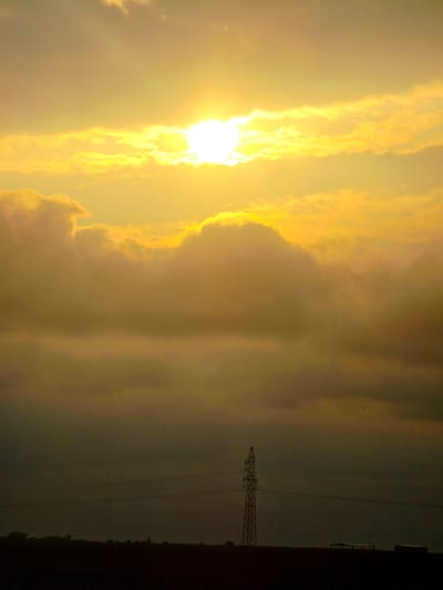 A dramatic sunset is captured in Brzezina, Poland, with the sun a brilliant orb peeking through thick, golden clouds, casting an ethereal glow over the scene. The lower half of the image is dominated by a dark, silhouetted landscape. A tall, lattice-style electricity pylon stands prominently against the dusky sky, its lines stretching horizontally across the frame. In the distance, the faint outlines of trees and structures, including a small church spire, are visible on the horizon. The overall atmosphere is one of serene twilight, transitioning into night.