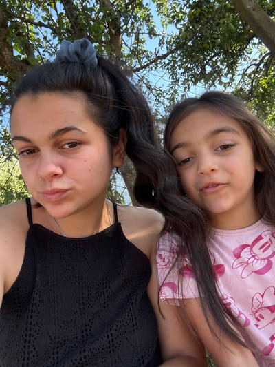 The media file is a selfie taken outdoors, featuring a woman and a young girl positioned closely together. The woman, in the foreground, has dark hair pulled back with a grey scrunchie and is wearing a black, lacy top with a silver necklace. The young girl, positioned behind and above the woman, has long dark hair and is wearing a pink t-shirt with a repeating Minnie Mouse pattern.
The setting appears to be a natural environment with trees and blue sky visible in the background. The lighting suggests it is daytime, likely sunny. The woman's expression is relatively neutral, while the girl is smiling. The image is oriented vertically, with the subjects tilted. There is no visible text. The location context is Oreshak, Bulgaria.