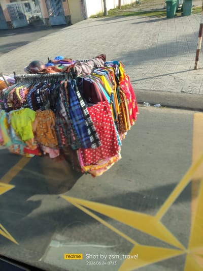 A street vendor's clothing rack is displayed outdoors on a paved sidewalk. The rack is filled with an assortment of colorful garments, including shirts, dresses, and patterned fabrics, some of which are folded and piled on the lower racks. A dark-colored helmet rests on top of some of the clothing. In the background, a building with blue gates and motorcycles parked outside is visible, along with green trash bins and a sidewalk with a patterned tile design. The scene appears to be a typical street view in Trảng Bàng, Vietnam, with the yellow lines of a road visible in the foreground, suggesting the clothing rack is situated along a street.
