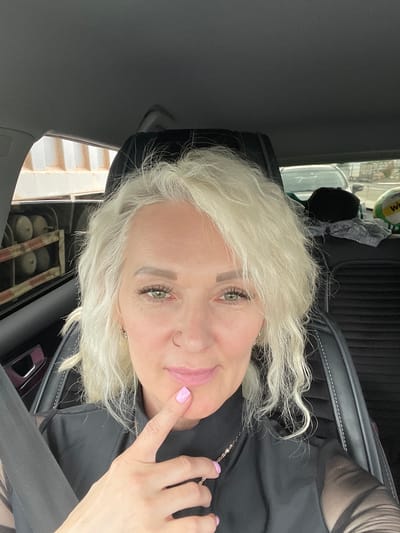 A woman with blonde, curly hair is sitting in the driver's seat of a car. She is wearing a black top with sheer sleeves and has a nose ring and a ring on her finger. Her left hand is raised, with her index finger touching her chin. Her nails are painted light pink. The car's interior is visible, including the headrest and seat. Outside the car, a truck bed with cylindrical objects is visible through the window, along with other vehicles and a construction site. The image was taken in Yam-Izhora, Russia.