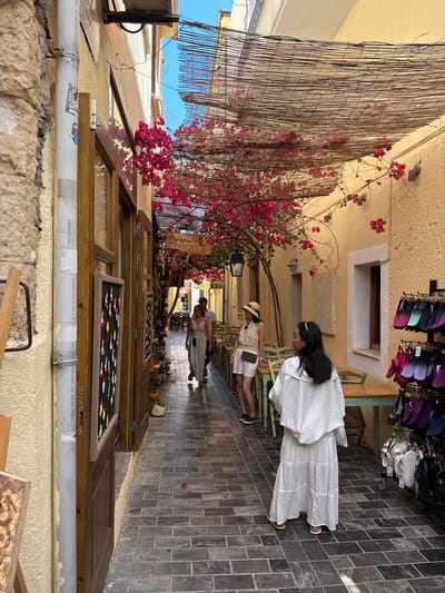 A narrow, cobblestone alleyway in the city of Rethymno, Greece, is lined with buildings adorned with blooming pink bougainvillea. A woven bamboo canopy shades the walkway. On the right, shelves display an array of colorful flip-flops and sandals. Tables and chairs are set up outside a building on the left, likely a cafe or restaurant. A woman wearing a white dress and headscarf stands facing away from the camera, while another woman in a straw hat and patterned top is visible further down the alley. The stone and yellow-painted walls of the buildings create a rustic atmosphere.
