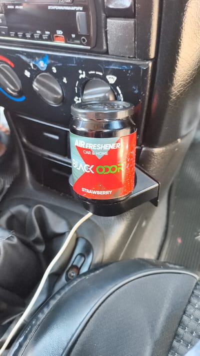 An air freshener is placed in a car's console in the city of Mirzo Tursunzoda, Tajikistan. The air freshener is a small glass jar with a red and green label that reads "AIR FRESHENER CAR & HOME BLACK ODOR STRAWBERRY". The car's dashboard is visible, with knobs and a radio. A white cable is plugged into the air freshener. The interior of the car appears to be well-used, with some wear and tear on the dashboard. The lighting is dim, suggesting it may be daytime or evening.