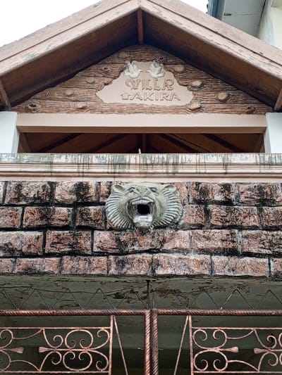 The image showcases a building facade with a rustic, textured aesthetic. "Villa Takira" is inscribed on a sign under the roof, adorned with two small bird figures. Below, a brick wall features a lion head sculpture with an open mouth. The gate in front of the wall is wrought iron with decorative swirls. The color scheme is dominated by earth tones, mainly brown, beige and hints of pink.
