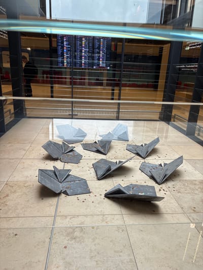 The image depicts an indoor scene at Berlin Brandenburg Airport (BER) in Schönefeld, Germany. The foreground features several metal origami-style sculptures placed on a tiled floor. Scattered around these sculptures are numerous coins, suggesting they might be part of a wishing well or donation setup. In the background, a departure information board displays flight details. A person wearing glasses and a backpack stands near the information board, looking at it. The overall environment appears to be a public space within an airport terminal, characterized by polished floors, modern architecture, and displays. A reflective glass barrier separates the viewer from the scene.