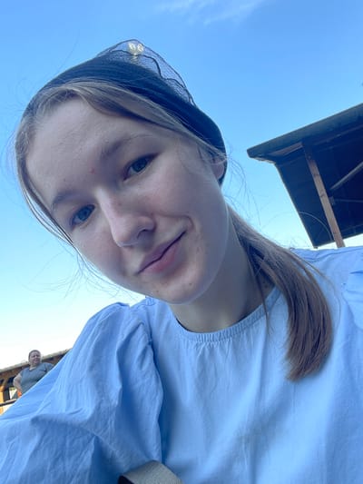 A young woman with light brown hair and a black head covering is taking a selfie outdoors. She is wearing a light blue ruffled shirt. In the background, there is a wooden structure, a clear blue sky, and another woman standing further away. The lighting suggests it is daytime. The exact location in Beltershain, Germany is not discernible from the image.