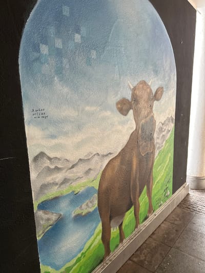 This image captures a vertical, arched mural painted on a textured wall within a covered public passageway or underpass in Schongau, Germany. The mural depicts a brown cow with small horns, facing forward, in an alpine landscape featuring a bright blue sky with subtle square patterns, distant grey mountains, and a blue lake with green surrounding terrain. Below the painted scene, a silver-colored metal trim borders the bottom edge of the mural, extending to the right. To the left of the cow, inscribed on the blue sky portion of the mural, is handwritten text in Cyrillic script that reads: "Я люблю Артёма но он пидог" (transliterated as "Ya lyublyu Artyoma no on pidog," meaning "I love Artem but he's a faggot"). The unpainted sections of the wall framing the mural are dark, and a light-colored fluted column is visible on the far right, indicating an architectural structure. The floor consists of light-colored square tiles on the left and darker, possibly patterned, tiles on the right.