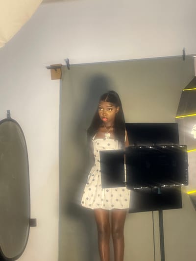 In this vertically oriented photograph, a young Black woman is the central subject, positioned slightly off-center and appearing to be looking at the camera. She has long, dark hair and is wearing a white dress adorned with dark polka dots. A noticeable detail is a shiny, possibly diamond-encrusted necklace around her neck. Her expression could be interpreted as playful or seductive, with her tongue slightly sticking out.
The setting appears to be a studio, indicated by the presence of what look like professional lighting equipment and a backdrop. The background consists of a light gray surface, possibly a wall or a fabric backdrop. To the left of the woman, there's a glimpse of a soft, light-colored object, potentially a piece of furniture or another studio prop. At the bottom of the image, a large, dark, oval-shaped object is visible, which could be a reflector or a large camera lens.
Above the woman, several rectangular, black objects are arranged, likely part of the lighting setup. The lighting in the scene is somewhat dramatic, with highlights and shadows suggesting artificial illumination. The overall mood is suggestive and artistic. There is no visible text in the image. The time of day and weather are not discernible from the image. Given the location context of South Ropp, Nigeria, the image captures a fashion or portrait photography session.