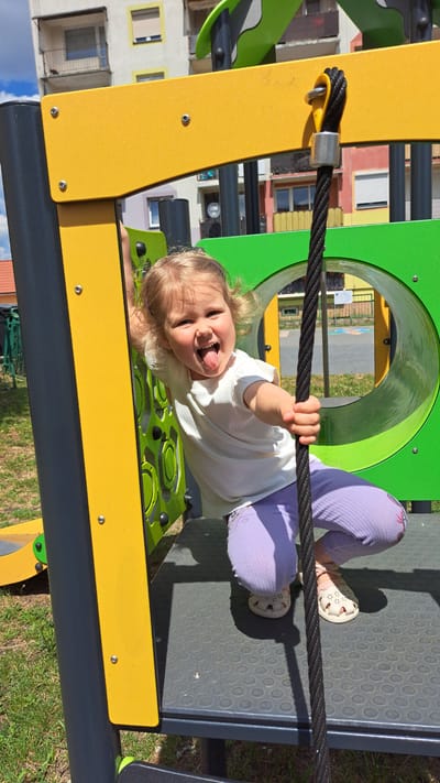 In Krosno Odrzańskie, Poland, a young Caucasian girl is posing on a playground. She is squatting, holding onto a thick black rope, and sticking out her tongue. She is wearing a white t-shirt and light purple leggings. The playground equipment is brightly colored with yellow, green, and grey sections. Behind the girl, a circular green tunnel is visible. The scene is set outdoors with green grass and a building in the background under a bright sky.