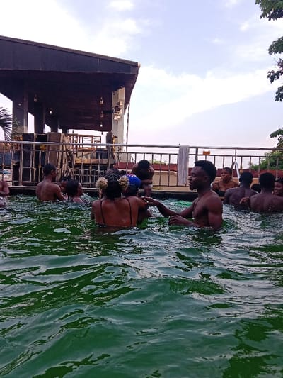 The image captures an outdoor scene at a swimming pool, likely during the late afternoon or early evening given the soft, diffused light and the appearance of the sky. Numerous people are in the pool, predominantly young adults, engaged in what appears to be a social and recreational gathering.

In the foreground and mid-ground, the rippling surface of the water dominates the lower half of the frame. The water is a distinct deep green color, suggesting it might be a natural spring pool, or perhaps an unchlorinated pool with a build-up of algae, rather than a typical blue chlorinated pool.

Several individuals are partially submerged in the water, with most visible from the chest or shoulders up. On the right side of the mid-ground, a man with a short beard and dark hair is smiling and looking towards another person to his left. He is bare-chested and wears a necklace. His right hand appears to be gently touching the arm or shoulder of a woman next to him. This woman is facing away from the camera, wearing a dark-colored swim top with thin straps. Her hair is styled with two decorative, light-colored puffs or buns on the sides of her head. To her left, another person is also visible from the back.

Further into the pool, closer to the background railing, more people are present. Many have their backs to the camera, while others are partially obscured. They seem to be standing or wading in the water, interacting with each other.

The background features a structure with an extended, dark-colored roof supported by pillars, possibly part of a poolside bar, restaurant, or changing facility. Several pendant lights hang from beneath this roof. A metal railing runs horizontally across the mid-background, separating the pool area from this structure. A white towel is draped over this railing towards the center-right. Above the building and railing, a bright, cloudy sky is visible. On the far right, some green foliage or trees can be seen.

The overall atmosphere of the image is one of leisure, enjoyment, and social interaction among the group in the pool. No discernible text is visible anywhere in the image.