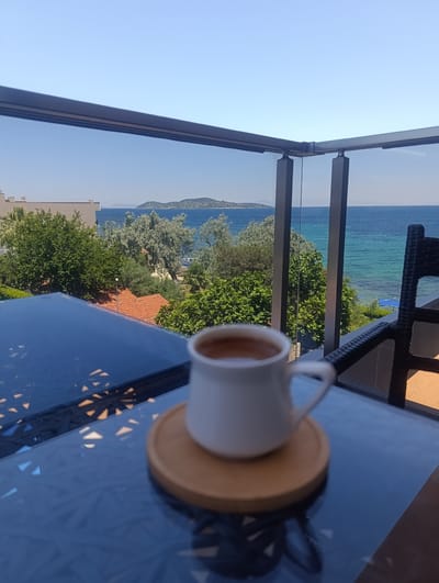 A white mug of coffee sits on a glass-topped table on a balcony, overlooking a serene seascape. The coffee is a rich brown, with a frothy top. The mug rests on a small, round wooden coaster. Beyond the table, a glass railing separates the balcony from the view.

The scene is bathed in bright, natural light, suggesting it is daytime, likely mid-morning or afternoon, given the clear, vibrant blue sky with no visible clouds. The sea is a striking turquoise near the shore, transitioning to a deeper blue further out. In the distance, a chain of islands with green-covered hills dots the horizon.

Lush green trees and foliage fill the middle ground, interspersed with glimpses of red-tiled rooftops, indicating a coastal town or resort. A modern building with white walls is visible on the left side of the frame, partially obscured by trees. A dark wicker chair is positioned on the right side of the balcony, suggesting a place for relaxation.

The overall mood is peaceful and tranquil, evoking a sense of quiet enjoyment and relaxation. The reflections on the glass table add an artistic touch, with abstract patterns of light and shadow. No text is visible in the image. The location context suggests this is a balcony in Urla, Turkey, offering a beautiful view of the Aegean Sea.