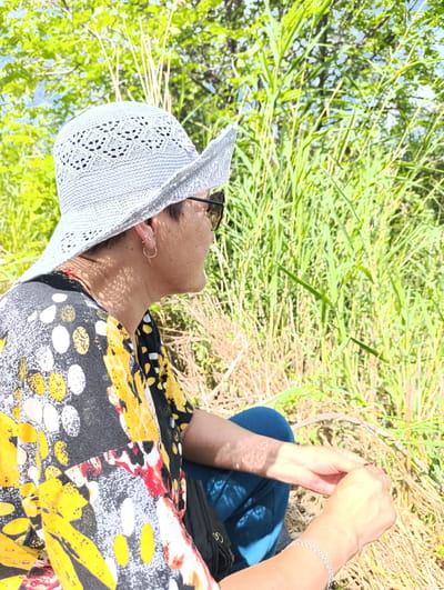 The image shows a person wearing a light blue crocheted hat and a colorful patterned shirt sitting outdoors. The person is facing to the right, with sunglasses on. Green vegetation, likely grass and trees, is visible in the background. The ground appears to be dry grass. The lighting suggests it is daytime, possibly sunny with some dappled shade from the foliage. The location is identified as the village of Zhdanov Rural District, Kazakhstan.