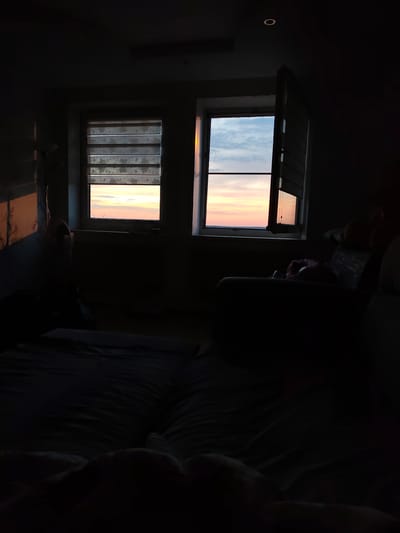 The interior of a darkened room is captured at dusk or dawn, as suggested by the colors visible through two windows. The window on the left has a partially drawn blind, revealing a sliver of the sky with hues of pink and orange. The window on the right is open, providing a clearer view of the sky's gradient of colors. A bed with rumpled covers occupies the foreground. To the right of the windows, an armchair or sofa is partially visible. The lighting is dim, with the natural light from the windows providing the main source of illumination. A single recessed light fixture can be seen on the ceiling. The image does not show any people. The location is not discernable as Gremyachinsk, Russia based on the image alone.