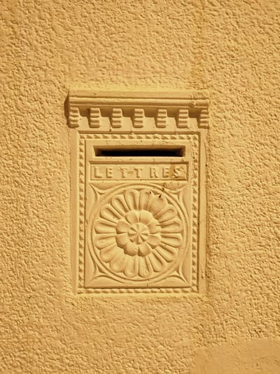 An antique, recessed mailbox is set into a textured, ochre-colored wall in Ariana, Tunisia. The mailbox itself is made of a lighter, sandy-colored material and features decorative elements. Above the mail slot, the word "LETTRES" is inscribed. Below the slot, a circular, floral motif is carved into the surface. The mail slot is a dark, rectangular opening. The wall has a rough, stucco-like finish. The lighting suggests a sunny day, casting shadows that highlight the texture of the wall and the relief of the mailbox's decorations.