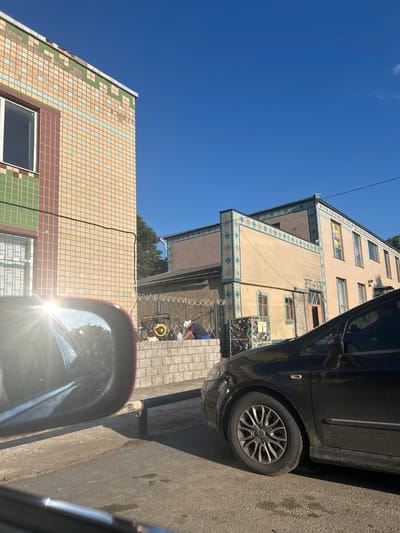 This image depicts an outdoor scene in Konopliane, Ukraine, on a clear, sunny day. The perspective is from inside a vehicle, looking outwards and upwards. Dominating the frame are two buildings, one to the left and a taller one to the right, both clad in patterned tiles. The building on the left features cream and green tiles, with a visible window. The taller building on the right has cream tiles with decorative blue and pink accents on its lower levels, and several windows visible.

Parked alongside the taller building is a dark-colored car, its front wheel and part of its body clearly visible. The car is positioned on a paved road. Further down the street, near the base of the taller building, a yellow generator is visible, connected to electrical wires. A person, wearing a blue shirt and dark shorts, is standing near the generator, seemingly engaged in some activity.

The scene suggests it is daytime, likely late morning or early afternoon, given the bright sunlight and the harsh shadows cast. The clear blue sky indicates fair weather. The presence of the buildings, the car, and the generator suggest an urban or semi-urban residential or commercial area. There is no discernible text visible in the image. The overall mood is one of everyday activity under bright daylight conditions.