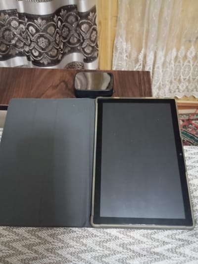 An open tablet computer with a dark gray cover is placed on a textured surface. The tablet screen is black and is on the right side of the image. The cover is on the left side of the image. In the background, there is a wooden table and curtains. A small black object with a reflective surface, possibly a mirror, is also on the table. The overall scene appears to be indoors.