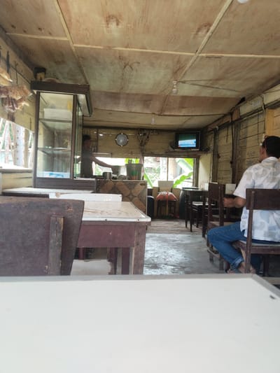 The image depicts the interior of a modest eatery, likely located in Keudegeudong, Indonesia. The space is enclosed with wooden walls and ceiling, and is lit by at least two visible light bulbs. A man, wearing a white and grey patterned shirt and blue jeans, is seated on a wooden chair at one of the tables. A second person, possibly a vendor, is standing behind a counter near a glass display case. A television is mounted on the wall near a window, showing a broadcast. Other wooden chairs and tables are arranged within the space, and various objects, including bags and containers, are placed near the windows. The visible tables have white tops, and the floor is concrete.