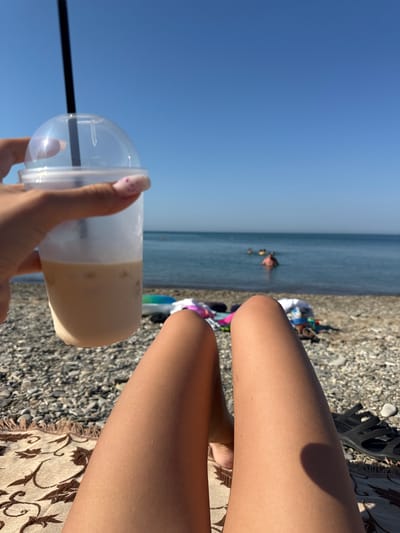 The image shows a close-up view of a person's legs and a drink on a pebble beach, with the sea and sky in the background. The person is lying on a patterned blanket, and their legs are tanned. They are holding a clear plastic cup filled with a light-colored beverage, possibly an iced coffee or latte, with ice cubes visible. A black straw extends from the cup. In the distance, several people are visible in the calm, blue sea. The sky is clear and bright blue. The beach is covered in smooth, grey pebbles. The overall scene depicts a relaxing day at the beach. The location is specified as the city of Dzhubga, Russia.