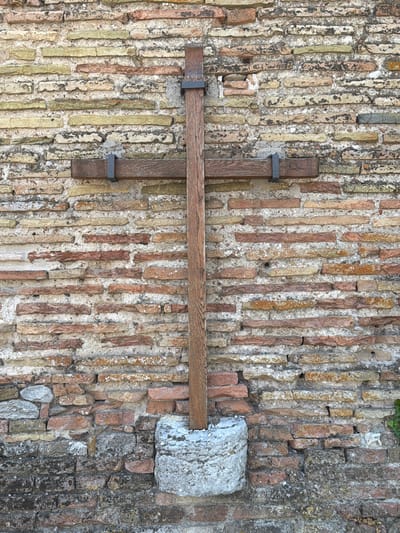 A wooden cross stands upright against a weathered brick wall. The cross is made of dark wood and is secured to the wall with metal brackets. The brick wall is constructed of various shades of red, orange, and yellow bricks, some of which are chipped or broken, with visible mortar. The cross is supported at its base by a rough, cylindrical stone. The scene appears to be outdoors, with hints of greenery on the right side of the image. This image was taken in Santarcangelo di Romagna, Italy.