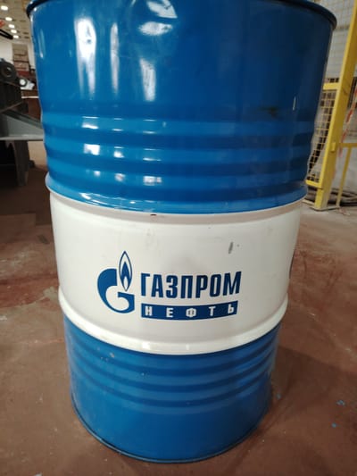 The image shows a blue and white metal barrel with the Gazprom logo and text "НЕФТЬ" (Nefte, meaning Oil) printed on its white section. The barrel is positioned in what appears to be an industrial or storage area, with a reddish-brown floor and some machinery visible in the background on the left. A yellow metal structure, possibly a safety railing or part of equipment, is on the right. The lighting suggests an indoor environment. The image is a close-up, centered on the barrel. There are no people or animals present in the frame. The scene appears to be in a location within Краснадворскі сельскі Савет, Belarus.