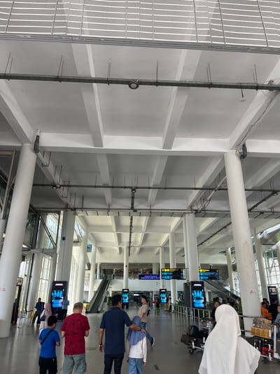 The image depicts the interior of an airport terminal in Ramunia Dua, Indonesia. The high ceiling is structured with a grid of white beams and pillars. Metal conduits and wires are visible along the ceiling and walls. People are present, some walking, some standing, and some with luggage. There are signs indicating directions and information displays showing flight details or advertisements. An escalator is visible in the background. The floor is tiled and appears clean. Natural light enters through glass windows.