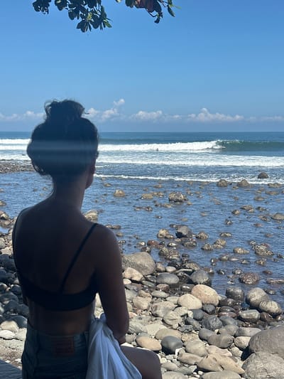 The image shows a person standing on a rocky beach in Pekutatan, Indonesia. They are facing the ocean, which has moderate waves. The person has dark hair tied up in a bun, is wearing a dark-colored top and denim shorts, and has a white shirt draped over their shoulder. The beach consists of numerous smooth, dark-colored rocks of varying sizes. To the left of the frame, foliage from a tree is visible. The sky is a clear blue, with some scattered clouds near the horizon.