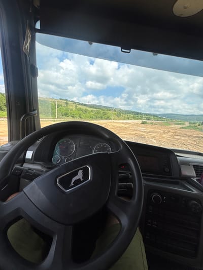 An internal view from the driver's perspective inside a heavy vehicle, likely a MAN truck, overlooking a vast outdoor landscape in Verinsko, Bulgaria. The image is captured during the daytime under a bright, partly cloudy sky.
In the foreground, a black steering wheel with a distinctive silver stylized lion logo (identifying it as a MAN vehicle) dominates the lower right. Through the steering wheel, the instrument cluster is visible, displaying gauges for RPM (marked up to x100 RPM), speed (up to 100 km/h), fuel level, and engine temperature. A small digital display area within the cluster shows some Cyrillic text, including what appears to be "СООБЩЕНИЕ" (message) and "ОШИБКИ" (errors), suggesting informational or warning indicators. Below the dashboard, a leg clad in olive-green trousers is partially visible, indicating a person is seated in the driver's position.
To the right of the steering wheel, a section of the central console features climate control dials and buttons, including a large 'C' for temperature, and what appears to be a turned-off infotainment screen.
Through the large windshield, a wide panorama of the outside environment unfolds. The immediate area outside the vehicle is characterized by a significant expanse of light brown, disturbed earth, possibly indicating a construction site, quarry, or agricultural land under preparation. Beyond this, patches of green vegetation and trees stretch towards a horizon line marked by distant hills or mountains. Utility poles are visible across the landscape. Overhead, a blue sky with scattered white clouds occupies the upper portion of the view. A black sun visor is visible along the top edge of the windshield frame, and a black grab handle is mounted on the driver's side A-pillar.
The scene suggests the vehicle is either parked or being driven through a rugged, open-air work environment.
