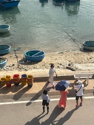 This overhead shot depicts a vibrant waterfront scene in Ninh Chu Ward, Vietnam, under strong sunlight. The image is divided into three main sections: water, a sandy embankment, and a paved area/road.

On the left, calm, grey-green water is visible, featuring several traditional Vietnamese basket boats (thúng chai) that are either partially submerged or moored. Ropes stretch from these boats towards the shore. The middle section is a sandy embankment, noticeably cluttered with a variety of debris, including plastic bottles, bags, and discarded sandals. One blue basket boat is resting on this sandy stretch.

To the right, a paved walkway or road runs parallel to the water. Four people are present in this area, casting long shadows indicative of either morning or late afternoon sunlight. One person, wearing a white shirt, black shorts, and a white cap, is holding a smartphone as if taking a photograph. Nearby, another individual in a long, reddish-pink dress is holding a dark blue umbrella open, likely for protection from the sun. Further along, a person in a light-colored long dress is visible, and in the foreground, another individual in a grey top and black shorts/leggings walks. Adjacent to the paved area, a stack of colorful plastic buckets or baskets is neatly arranged. A white concrete bench or railing also lines the paved edge.

The scene captures a blend of daily life and local industry, with the basket boats and stacked baskets suggesting fishing or market activities. No discernible text is visible.