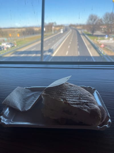 The image, correctly oriented, displays an indoor setting looking out onto a bustling multi-lane highway in broad daylight. On the left, a large window reveals an autostrada under a clear, bright blue sky, with the sun casting strong light on the road surface and surrounding landscape. Vehicles are visible along the highway, some in motion and others seemingly parked at a distant service area or rest stop. The window itself shows some smudges or reflections.
On the right side of the image, indoors, a piece of food resembling a large sandwich, possibly made with focaccia or a similar bread, rests on a white rectangular tray. The food is partially wrapped in a light brown paper napkin, on which the word "EATALY" is clearly visible. The tray and food are placed on a dark-colored surface, likely a table or windowsill. No people are visible in the frame.
The scene suggests a moment of rest, with someone having a meal or snack while observing the traffic on a major road. The clear weather indicates a sunny day. The prominent "EATALY" branding on the napkin, combined with the characteristics of the highway, strongly points to a location in Italy. This aligns with the provided context of Dorno, Italy, a commune typically situated near such major transportation routes.