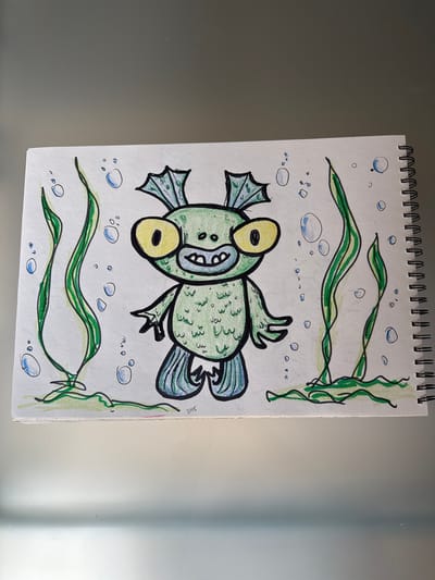 This is a close-up, top-down shot of a page from a spiral-bound sketchbook. The sketch depicts a cartoonish green fish with large yellow eyes, a wide grin, and fins. The fish is surrounded by blue bubbles and green, wavy seaweed, suggesting an underwater scene. The drawing is rendered with black ink outlines and colored with green, blue, and yellow. The sketchbook page is white and slightly textured. The background of the image is a reflective, grey surface, possibly a table or a wall, with a faint vertical highlight. There is some visible text on the right side of the drawing, "SOLL," written vertically. The lighting is bright and even, casting subtle shadows. The context of Fañabé, Spain, is noted, but there are no visual cues in the image to confirm this specific location.