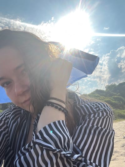 A young person with brown hair is sitting on a beach, looking at the camera. They are wearing a black and white striped shirt. A blue umbrella is visible behind them, and the sun is shining brightly in the sky, creating lens flare. The background shows green trees and blue sky with clouds. The sand is visible in the foreground. The location is Ahtopol, Bulgaria.