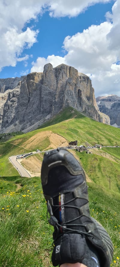 A first-person perspective photograph captures a black hiking boot in the foreground, positioned on a grassy slope dotted with small yellow wildflowers. The boot is laced with a quick-lace system, and a stripe of red, yellow, and blue is visible on its side. Behind the boot, a winding asphalt road curves through a lush green landscape, bordered by parked cars. Several buildings, appearing to be a hotel and associated structures, are situated on a ridge overlooking the road. In the background, a massive, imposing mountain range with steep, rocky faces dominates the horizon under a bright blue sky with scattered white clouds. The overall scene depicts a scenic mountain vista in the Italian Dolomites, likely within the Sëlva - Wolkenstein - Selva di Val Gardena area, suggesting a hiking or outdoor recreation setting.