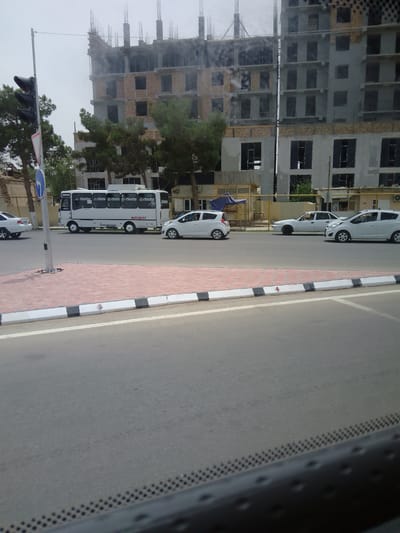 The image captures a street scene in Termez, Uzbekistan, with a multi-story building under construction in the background. The building features exposed brickwork and unfinished floors, with rebar protruding from the top. In the mid-ground, a white bus with "ISUZU" branding and "INTURIST" text is parked or driving slowly. Several white passenger vehicles are visible on the road. A traffic light stands on the left side of the road, next to a blue directional sign. The foreground shows a paved road with a sidewalk featuring red and white brickwork and a black and white striped curb. The sky is overcast.