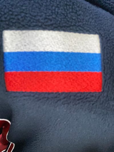 The image shows a close-up of a stitched patch depicting the Russian flag. The flag has three horizontal stripes: white on top, blue in the middle, and red on the bottom. The flag is attached to a dark blue fabric, possibly part of a garment or textile. There are no people or any environmental context visible that would indicate a specific location in Luzhitsy, Russia. The focus is solely on the flag patch.