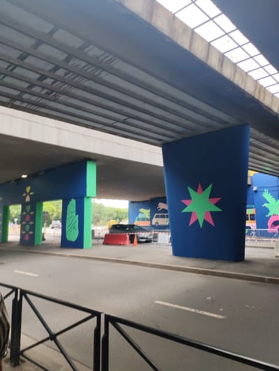 The image captures an urban underpass in Fontenay-sous-Bois, France, from a ground-level perspective. The scene features a concrete bridge structure overhead, supported by multiple pillars. The underside of the bridge exhibits horizontal concrete beams with visible weathering and dark streaks. A grid of translucent or light-emitting panels is present on the upper right section of the bridge's ceiling.

The pillars and lower walls of the underpass are painted with vibrant murals. A large, broad blue pillar on the right displays a prominent two-toned star design, consisting of a green star superimposed on a pink star. To the left, a blue wall section features a large green open handprint. Further left, another blue section is adorned with a repeating pattern of stylized green and pink floral or leaf shapes. The structural supports for these painted sections are colored bright green.

An asphalt road runs through the mid-ground, marked by a single white dashed line. On the right side of the road, a dark-colored SUV is parked, positioned behind a red concrete barrier. An orange traffic cone is visible near the barrier and the SUV. White temporary metal fences are also present behind these objects. In the immediate foreground on the left, a black metal railing with X-shaped supports is partially visible. Beyond the underpass, the background reveals an open area with green foliage, consistent with a park or natural space, under an overcast sky. No individuals or specific actions are discernible within the frame.