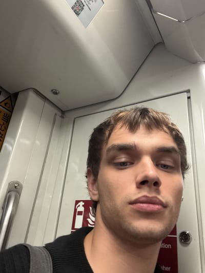 A medium shot captures a young Caucasian male looking directly at the camera, his face filling the majority of the frame. He has light brown hair styled with bangs that fall across his forehead, and blue eyes. His lips are slightly parted, and he appears to be in a public transportation setting, likely a subway car.
The background shows the interior of the subway car, with white walls and paneling. A chrome handrail is visible on the left side of the image. A red and white fire extinguisher symbol is affixed to the wall on the right. Above the man's head, a sticker with a QR code and Chinese characters is partially visible on the ceiling. The overall lighting is bright and artificial, typical of indoor public spaces. The image suggests the man is taking a selfie while traveling. The context of Changning District, China is not evident from the visual information alone.