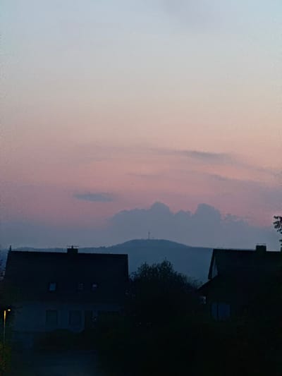 A dimly lit evening sky, transitioning from pale blue to soft pink, hovers above a silhouetted landscape. In the foreground, the dark outlines of houses with visible roof antennas and chimneys are prominent. A streetlamp casts a faint glow. Further in the distance, a rounded hill is visible, topped with a tall, slender tower, likely a broadcast antenna. The overall impression is of a quiet residential area at dusk, with the tower serving as a distant landmark.