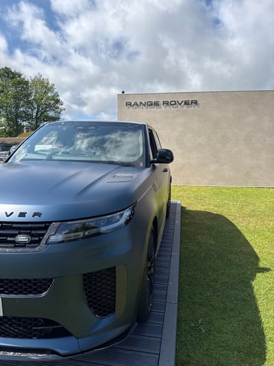 The image shows a dark blue Range Rover parked in front of a tan building in Bramham, United Kingdom. The building has the words "Range Rover" written on the side. The car is parked on a black and grey concrete surface. The car is facing the camera and a small portion of the front end of the car is visible. The car is surrounded by green grass. There is a blue sky with white clouds above. There is no one in the image.