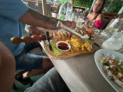 The image captures an outdoor dining scene at a restaurant in Kremen, Bulgaria. In the foreground, a person wearing a blue t-shirt and denim shorts is seated at a table, holding a skewer with grilled meat and vegetables. French fries and dipping sauces are served on a wooden board. To the right, another person is visible, wearing a bright pink off-the-shoulder top, with a glass of white wine in front of them. The table is set with plates, glasses, and a jug of milk or cream. The background reveals a restaurant setting with tables, chairs, and lush greenery, suggesting a relaxed outdoor ambiance. The overall scene depicts a meal being enjoyed in a restaurant environment.