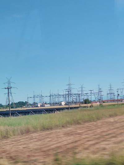 An outdoor, medium-long shot captures a vast electrical substation with numerous tall, metal towers supporting overhead power lines. In the foreground, a field of dry, brown grass is visible, partially blurred due to motion. Behind the substation, a line of green trees forms a backdrop against a clear, bright blue sky. The scene is located in Taxiarches, Greece, and depicts an industrial power infrastructure amidst a rural landscape.