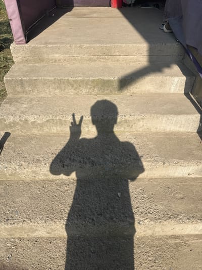 The image shows a shadow of a person making a peace sign on a concrete staircase. The staircase is outdoors, with grass and some purple-colored siding visible on the left. A shadow from an overhead structure casts a diagonal line across the stairs. The location is described as International, Kyrgyzstan, though this specific detail is not visually confirmed by the image content itself, which appears to be a generic outdoor setting.