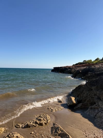 This is a vertical, eye-level, wide shot of a rugged coastline with a beach and the sea. The sky is a clear, cloudless blue, suggesting a bright, sunny day, likely late morning or early afternoon given the strong shadows. The sea is a tranquil turquoise, with gentle waves lapping at the shore. The beach is a mix of sand and pebbles, with several large, porous rocks scattered across it. A dramatic cliff face, covered in sparse vegetation at its peak, juts out into the sea on the right side of the frame. The textures of the rock are rough and pitted, with some areas appearing dark and damp from the water. The overall scene evokes a sense of peace and natural beauty. There is no visible text.