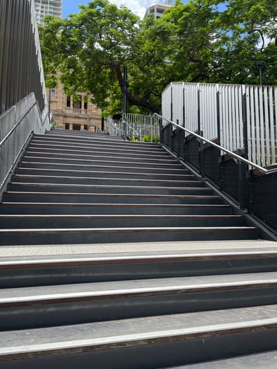 The image displays an outdoor urban scene in Brisbane City, centered on a modern, wide staircase or sloped walkway. The walkway is constructed from numerous parallel panels, alternating in color between dark grey and a lighter off-white or light grey, creating a distinct striped pattern. A sleek metal handrail runs alongside this structure.

To the left of the walkway, a large, lush green tree with dense foliage dominates the midground, its branches extending across a significant portion of the frame. Beyond the tree, glimpses of various urban buildings are visible, including one with a more traditional or older architectural style and another more contemporary high-rise. The sky above is a bright blue with a few scattered white clouds, indicating a clear and sunny day. The natural sunlight illuminates the scene, casting subtle shadows and highlights on the walkway panels, suggesting a time of day likely between late morning and early afternoon.

No people are present in the image, contributing to a sense of tranquility within the urban environment. The blend of modern infrastructure, established green spaces with mature trees, and diverse architectural styles are consistent with the urban landscape of Brisbane, Australia. There is no visible text in the image.