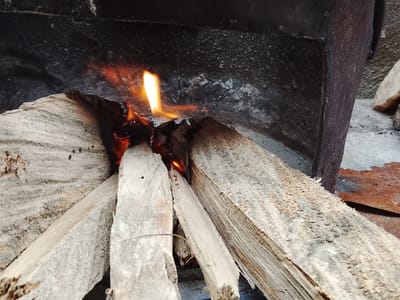 A close-up, low-angle shot captures a fire burning within a crude metal brazier, located outdoors on a paved surface, possibly in Ozuri, Nigeria. The brazier appears to be constructed from a dark, weathered metal, with visible signs of rust and wear. Stacked within the brazier are several pieces of light-colored, rough-hewn wood, some of which are actively burning. Flames, predominantly orange and yellow, are visible, igniting the edges of the wood. The background consists of the interior wall of the brazier, which is black and textured, and a portion of the outer wall on the right side, which is reddish-brown with visible rust. To the right of the brazier, a rough stone or concrete surface is visible, with a few scattered rocks. The lighting suggests daylight, with the fire being the primary light source in the immediate foreground. No people are present in the scene.