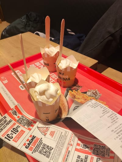 A red tray holds four KFC My Mini Sundaes, each topped with white ice cream and a wooden spoon. Beside the desserts is a piece of fried chicken and a bun. A long receipt and a KFC promotional voucher are placed on the tray. The voucher advertises 1 euro equaling 10 points in a loyalty program and a chance to win a sundae by completing a customer satisfaction survey. The background features a wooden table and the legs of someone wearing dark-colored pants.