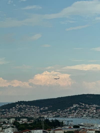 The image, oriented vertically, captures a panoramic view of a coastal town nestled against a verdant hillside, with a body of water dotted with boats in the foreground. The town is characterized by a dense cluster of buildings with red-tiled roofs, interspersed with white structures. The hillside is covered in dark green trees. In the sky, a small airplane is visible, appearing to fly between fluffy white clouds. The sky is a pale blue, with some areas of diffused light suggesting it might be late afternoon or early morning. The overall scene conveys a sense of tranquility and a picturesque Mediterranean setting, likely in Seget Donji, Croatia. There is no visible text in the image.