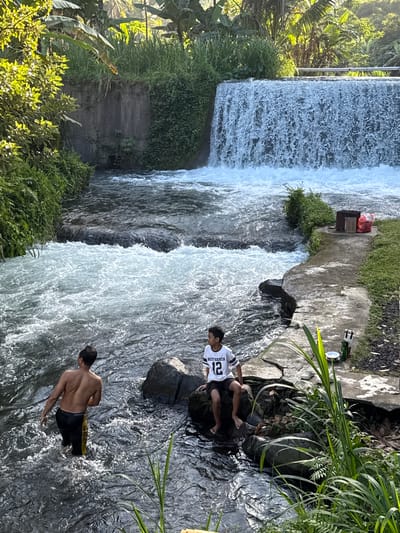 In Rendang, Indonesia, a natural setting features a flowing river with a small waterfall. Two boys are present. One boy stands in the river, facing away from the camera, wearing black shorts with a yellow stripe. The other boy sits on a rock in the river, wearing a white t-shirt with the number "12" and black shorts. The river flows from the waterfall, which is located in the upper right of the frame. The banks of the river are lined with lush green vegetation, including trees and tall grass. A concrete pathway runs along the right bank, and on it, there are two bags, one red and one brown, near a wire grill.