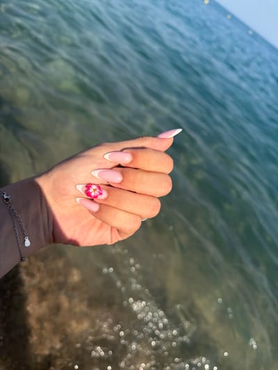 The image shows a close-up of a woman's hand with manicured nails, positioned against a backdrop of shimmering water and a sandy bottom. The nails are almond-shaped and feature a classic French manicure with a pink base and white tips. One nail is adorned with a small, vibrant pink and yellow flower design. A delicate silver bracelet with a small charm is visible on the woman's wrist. The water appears clear and shallow, with sunlight reflecting off its surface, creating a bokeh effect. The sandy bottom is visible beneath the water, with some darker patches and lighter sandy areas. The overall scene suggests a beach or coastal environment, consistent with locations in Santa Susanna, Spain.