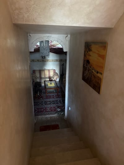 The image shows a view from the top of a staircase looking down into a room. The staircase is made of concrete and is illuminated by natural light. The room below is decorated in a traditional style, with a patterned rug, a sofa, and a coffee table. Above the sofa, there is a chandelier. A painting of animals in a savanna landscape hangs on the wall to the right of the staircase. The walls of the staircase and the room are painted in a light, earthy tone. The overall impression is one of an old, well-decorated house. The city of Yukki, Russia, is not visible in the image.