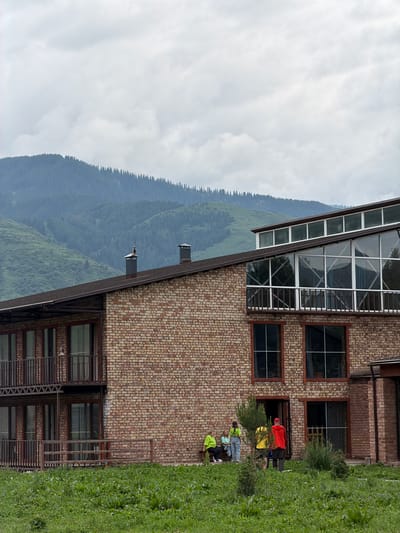 In Shabdan, Kyrgyzstan, a two-story brick building stands against a backdrop of verdant, rolling hills and a cloudy sky. The building features a mix of architectural styles, with a traditional brick facade on the lower floors and a modern glass-enclosed upper section. Several people are visible in the scene. On the ground floor, three individuals are seated on a bench, appearing to be in conversation, while two others stand nearby. A fourth person, wearing a bright red jacket, stands facing the building. The foreground is covered in lush green grass and vegetation, suggesting a natural or park-like setting surrounding the structure. The overall atmosphere is one of a serene, possibly recreational, location nestled within the Kyrgyz landscape.